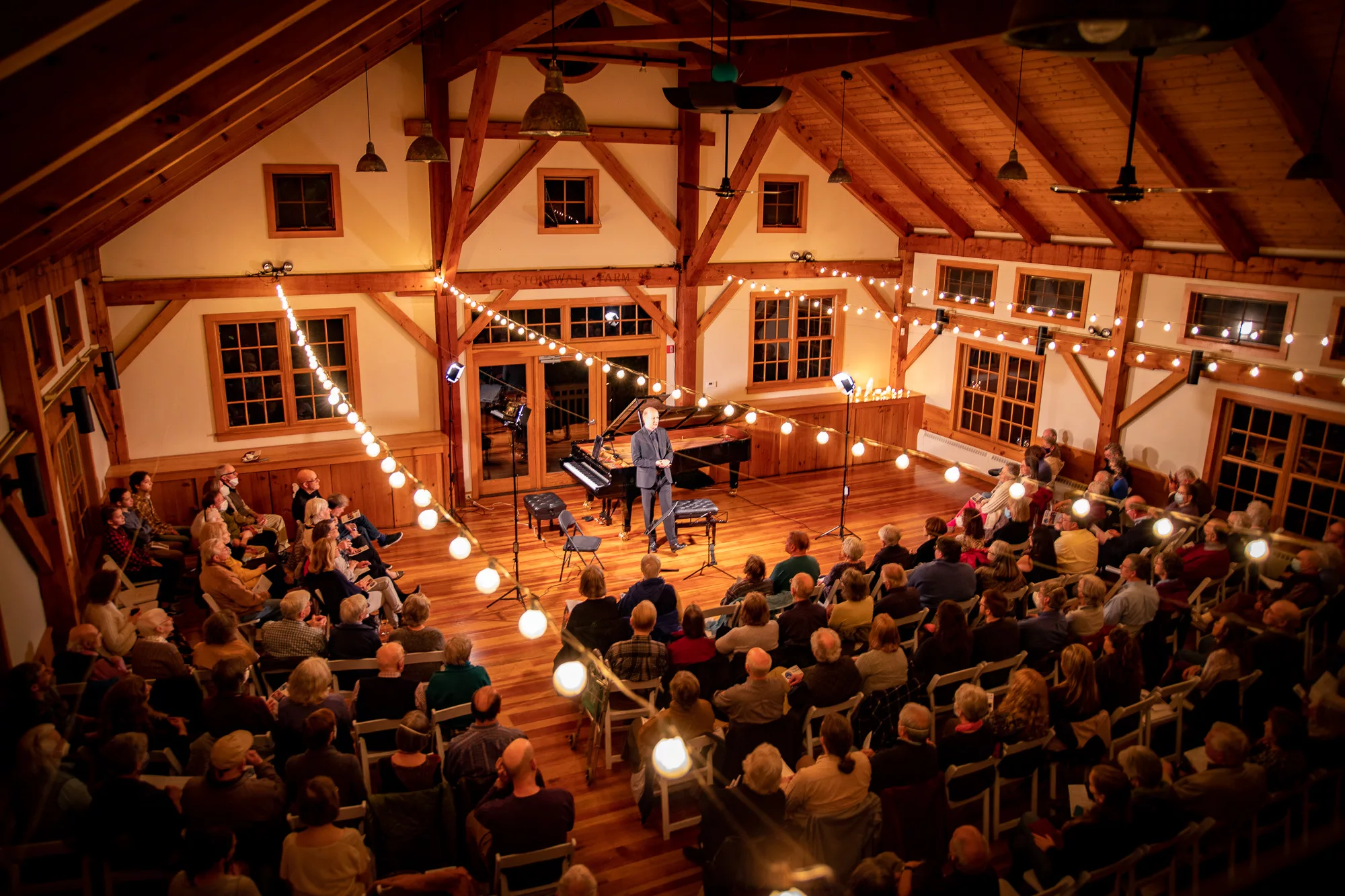 A warmly lit hall with audience members watching a stage performance beside a grand piano, under glowing lights. The cozy, attentive atmosphere invites you to enjoy all 11 concerts with the 2024-25 Full Season Ticket, including the full Winter Festival.