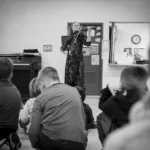 A woman plays the violin for attentive children seated on a classroom floor. The black-and-white scene evokes the joy of music—just like enjoying every performance with a 2024-25 Full Season Ticket (11 concerts, Winter Festival included).