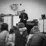 In a classroom, a man speaks to children seated on the floor while another adult smiles nearby. The scene promotes the 2024-25 Full Season Ticket: 11 concerts, including the full Winter Festival.
