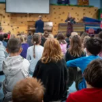 Children sit on the gym floor, attentively watching two adults present near a piano on stage, with gym equipment in the background—promoting the 2024-25 Full Season Ticket (11 concerts, includes full Winter Festival).