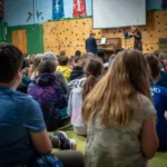 A group of children sit on a gym floor, facing a stage where a woman plays violin with a pianist beside her. The room features colorful banners, a climbing wall, and displays for the 2024-25 Full Season Ticket (11 concerts - includes full Winter Festival).
