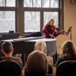 A woman plays violin and a man plays piano for an audience in a sunlit room—promoting the 2024-25 Full Season Ticket (11 concerts including the full Winter Festival).