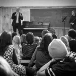 A man passionately addresses children in a classroom with a blackboard and piano behind him, as a woman stands nearby. The attentive group listens intently. The black and white photo promotes the 2024-25 Full Season Ticket (11 concerts, incl. Winter Festival).