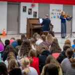 A group of children sit on the gym floor, watching a pianist and a violinist perform near posters promoting the 2024-25 Full Season Ticket (11 concerts including the full Winter Festival).