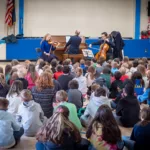 A group of children sits on a gym floor, watching musicians play piano, violin, and cello. Posters and a U.S. flag decorate the walls. Enjoy 11 concerts—including the full Winter Festival—with the 2024-25 Full Season Ticket.