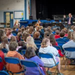 A man speaks to a large group of children seated on blue chairs in a gymnasium. Two musicians with string instruments sit at the front. The children face forward, listening attentively.