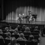 A black-and-white photo of a trio playing violin, cello, and piano on stage in front of an audience in a concert hall. The audience includes adults and children, some seated and one person in a wheelchair.