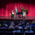 Three performers—a speaker at a podium, a woman with paper, and a cellist—are on stage with a grand piano and red curtain, as an audience looks on. Enjoy the experience live with the 2024-25 Full Season Ticket (11 concerts, incl. Winter Festival).
