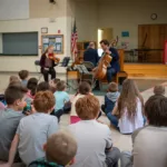 A group of children sit on a gym floor, watching two musicians play string instruments on a small stage in a school gymnasium. An American flag and colorful posters are visible in the background.