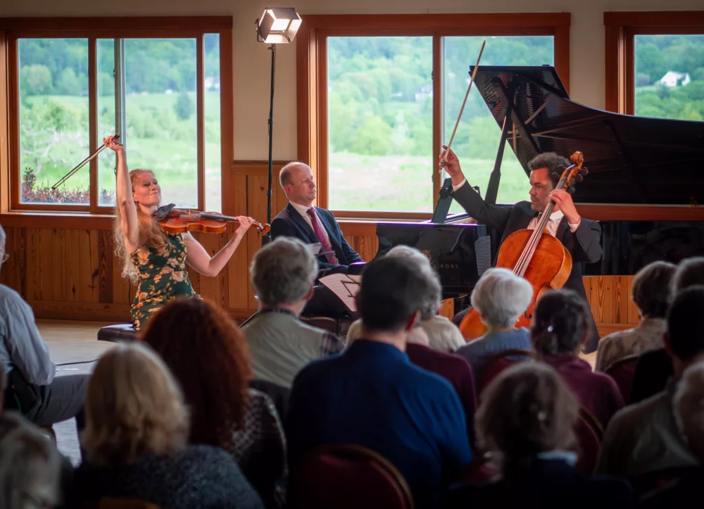 Musicians perform for an audience in a sunlit room with large windows showing green fields; a woman plays violin, a man plays cello, another man is at the piano—experience it all with the 2024-25 Full Season Ticket (11 concerts, incl. Winter Festival).