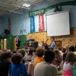 Children sit on a gym floor facing musicians performing on a small stage, with a speaker addressing the audience. Colorful banners reading “Effort,” “Kindness,” and “Knowledge” hang on the wall.