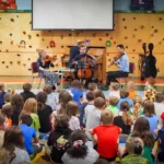 A string quartet performs for a group of children seated on the floor in a school gymnasium with a climbing wall in the background. The children watch attentively.