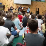 A large group of children sit on the floor of a gymnasium, facing musicians playing string instruments in front of them; colorful mats and a climbing wall are visible in the background.