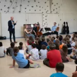 A group of children sit on the floor facing three musicians with string instruments and a man speaking to them in a gymnasium or multipurpose room, with a rock-climbing wall in the background.