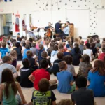 A large group of children sit on the floor in a gym, attentively watching a live musical performance by musicians playing string instruments and a piano at the front of the room.
