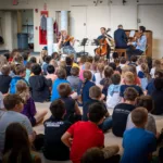 A group of children sit on the floor, facing a quartet of musicians playing string instruments and piano in a classroom or multipurpose room. The focus is on the attentive audience and the performing musicians.