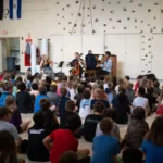 A large group of children sit on the floor facing a small classical music ensemble performing with a violin, cello, and piano in a gymnasium decorated with flags and a climbing wall.