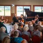 A classical string quartet performs in front of an attentive audience in a sunlit room with large windows and a grand piano, part of the 2024-25 Full Season Ticket (11 concerts, including the full Winter Festival).