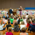 A group of young children sit on a gym floor, watching musicians perform live on stage decorated with colorful artwork—a taste of the experiences offered with a 2024-25 Full Season Ticket (11 concerts, including the full Winter Festival).