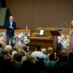 A woman plays violin on stage next to a piano, with a man standing nearby. Children watch from the audience in front of a colorful wall as part of the 2024-25 Full Season Ticket (11 concerts, including the full Winter Festival).
