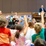 Children sit on a gym floor with hands raised, facing a man near a piano, as he presents in front of a colorful wall display—experience exciting moments like these with the 2024-25 Full Season Ticket (11 concerts incl. full Winter Festival).