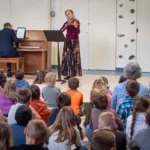 A woman violinist and a pianist perform for a group of attentive children in a school gym, capturing the spirit of live music celebrated with the 2024-25 Full Season Ticket (11 concerts including full Winter Festival).