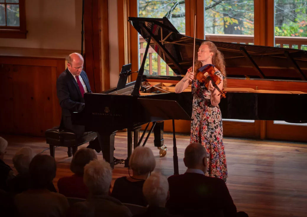 A woman in a floral dress plays violin beside a man in a suit at a grand piano on stage, performing for an audience with large windows behind—experience it live with the 2024-25 Full Season Ticket (11 concerts incl. Winter Festival).