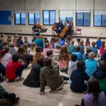 A group of children sits on a gym floor, attentively watching musicians with a cello, violin, and piano. The event promotes the 2024-25 Full Season Ticket (11 concerts – includes full Winter Festival), performed in front of large windows.