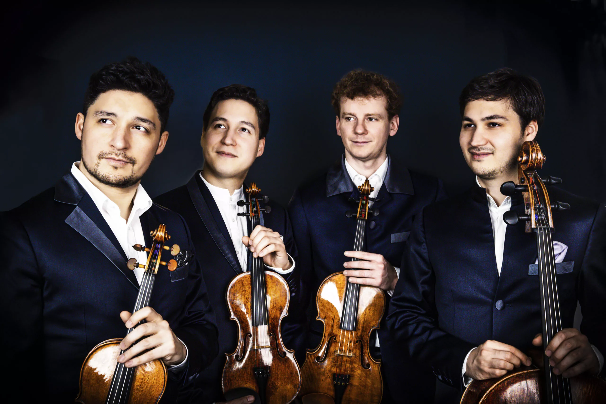 Four men in formal black suits stand together, each holding a string instrument (two violins, a viola, and a cello), against a dark background, looking slightly in different directions and smiling.