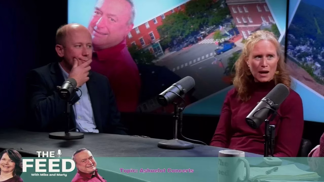 Two people sit at a podcast table with microphones, engaged in conversation. Behind them is a colorful background featuring photos of a man, a city street, and a building. Text at the bottom reads THE FEED - Topic: Ashuelot Concerts.
