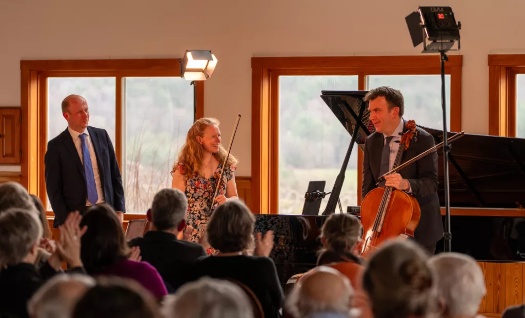 Three musicians stand in front of an applauding audience; one holds a violin, another a cello, and the third stands nearby. They are in a well-lit room with large windows and a grand piano in the background.