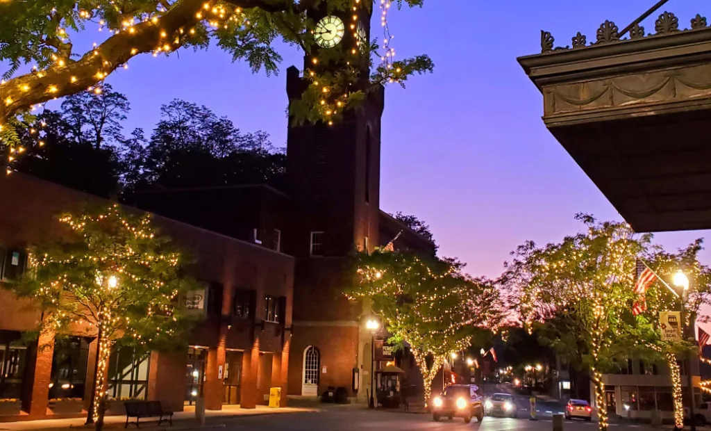 A small town street at dusk with trees wrapped in string lights, a brick clock tower, and a purple-orange sunset sky. Cars and streetlights line the road, creating a warm, welcoming atmosphere.