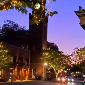 A small town street at dusk with trees wrapped in string lights, a brick clock tower, and a purple-orange sunset sky. Cars and streetlights line the road, creating a warm, welcoming atmosphere.