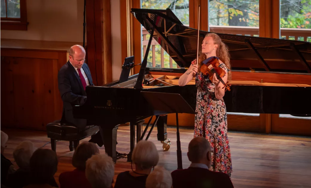 A woman in a floral dress plays violin while a man in a suit plays a grand piano on a wooden stage in front of an audience, with large windows and trees visible in the background.