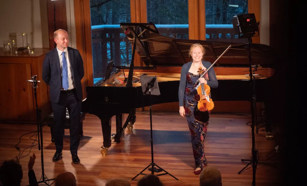 A woman holding a violin stands smiling next to a grand piano, while a man in a suit stands nearby. They are on a wooden stage with microphones and an audience in front of them.