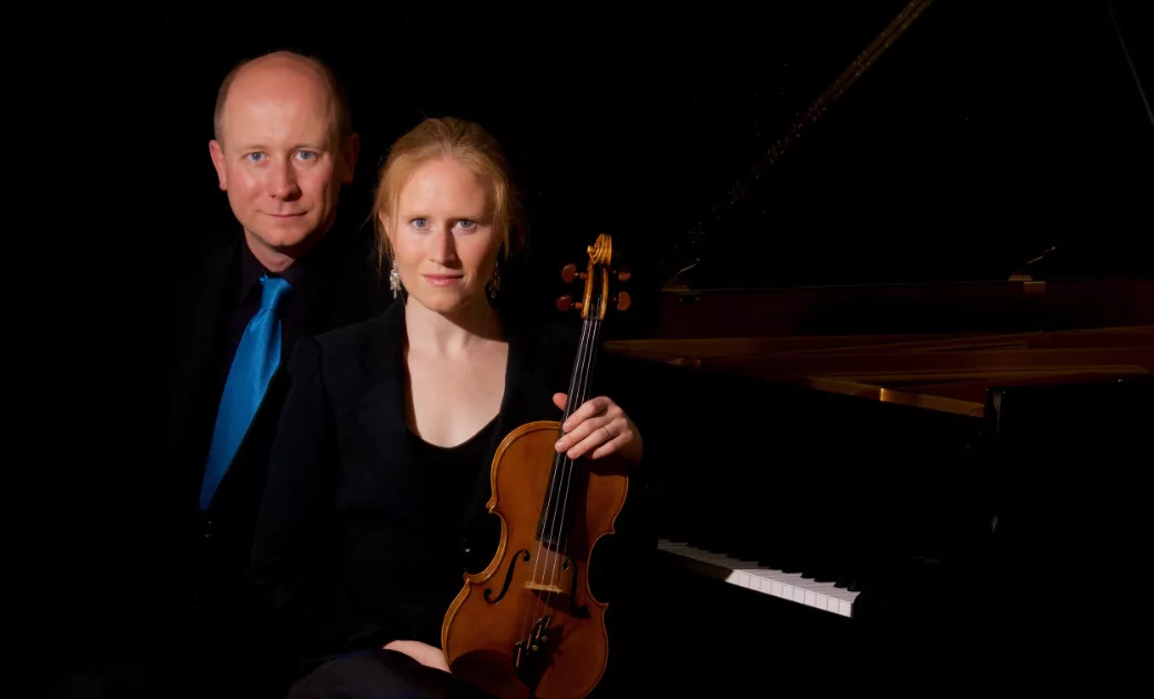 A man in a suit and blue tie stands behind a woman in black holding a violin beside a grand piano, both facing the camera against a dark background.