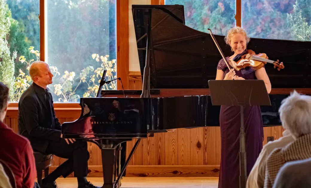 A woman in a purple dress plays the violin and smiles on stage beside a black grand piano, while a man in a suit sits at the piano. Audience members watch in a warmly lit room with large windows.