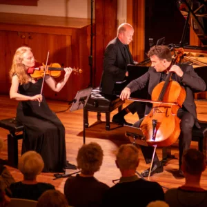 A trio of musicians perform on stage: a woman plays violin, a man plays cello, and another man plays piano. They are dressed formally, and an audience watches from the foreground in a wooden concert hall.