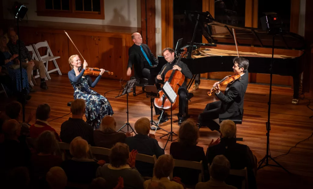Four musicians perform classical music on stage: a woman on violin, two men on cello and viola, and a man at the piano. An audience watches in a warmly lit wooden concert hall.