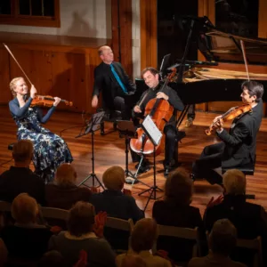 Four musicians perform classical music on stage: a woman on violin, two men on cello and viola, and a man at the piano. An audience watches in a warmly lit wooden concert hall.