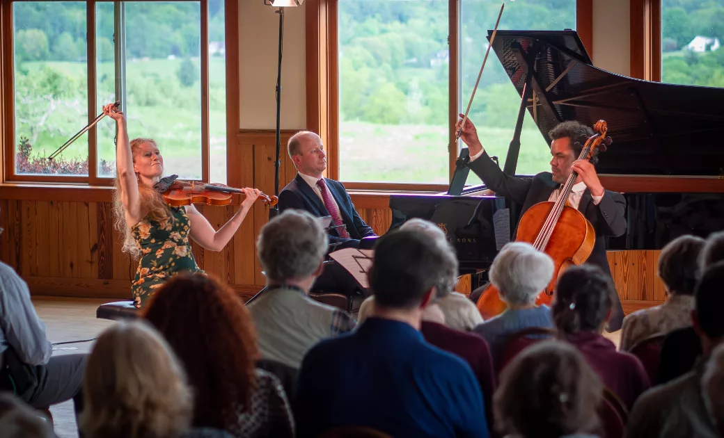 A violinist, pianist, and cellist perform together on stage in front of an audience in a light-filled room with large windows overlooking a green landscape. The musicians are passionately engaged with their instruments.