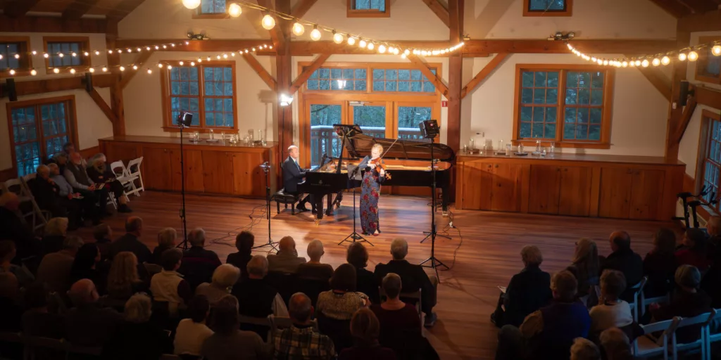 A violinist and pianist perform on stage in a cozy, wooden hall with string lights overhead, while an audience sits in rows, attentively watching the concert.
