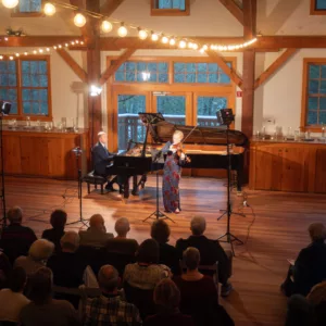 A violinist and pianist perform on stage in a cozy, wooden hall with string lights overhead, while an audience sits in rows, attentively watching the concert.