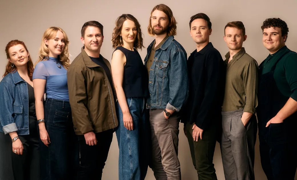 A group of eight adults stands in a row against a neutral background, smiling at the camera. They wear casual clothing in shades of blue, green, and gray.