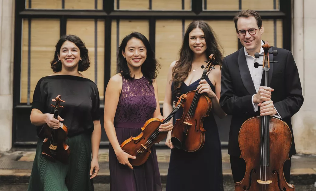 Four musicians stand together, smiling and holding string instruments—two violins, a viola, and a cello—against an outdoor background with a black-framed window and boarded-up panels.