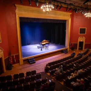 A few audience members with 2025-26 Bellows Falls Opera House Season Ticket - Premium Seating await the performance as a grand piano sits spotlighted on stage in a small, red-walled theater with ornate lighting.