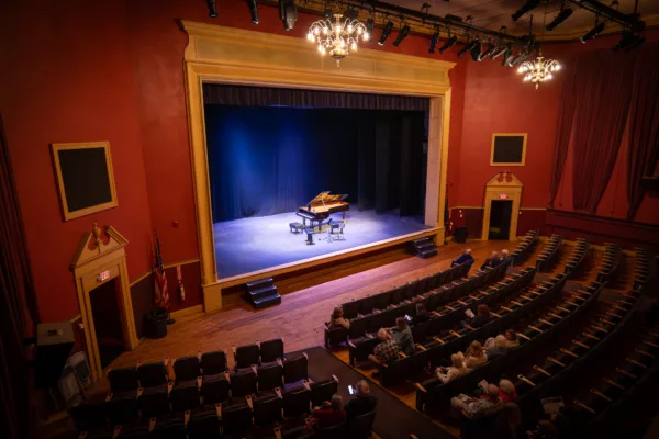 A few audience members with 2025-26 Bellows Falls Opera House Season Ticket - Premium Seating await the performance as a grand piano sits spotlighted on stage in a small, red-walled theater with ornate lighting.