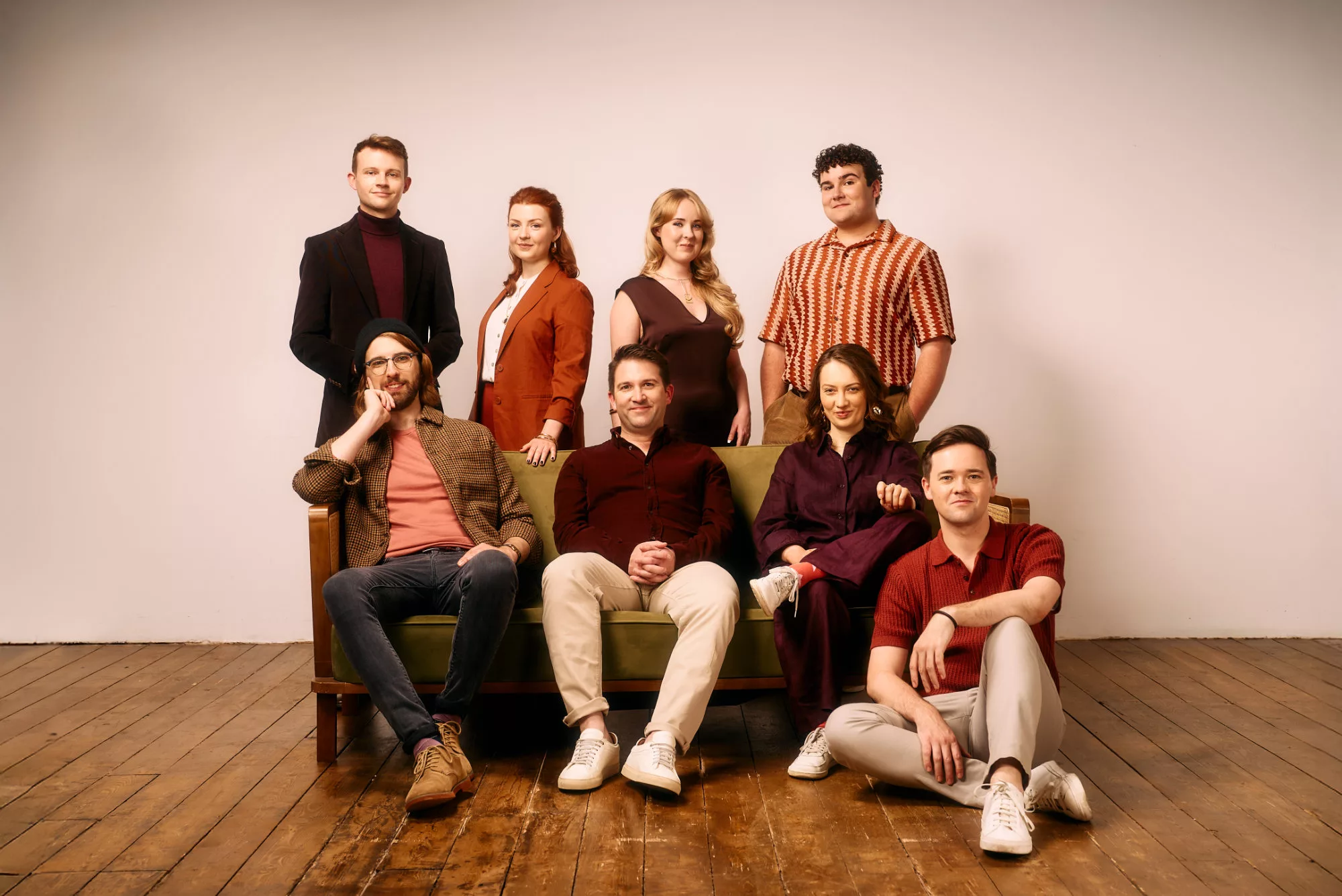 A group of eight adults posing for a photo in a studio with a wooden floor and plain backdrop. Five stand behind, while three sit on a green couch at the front. They are casually dressed and smiling at the camera.