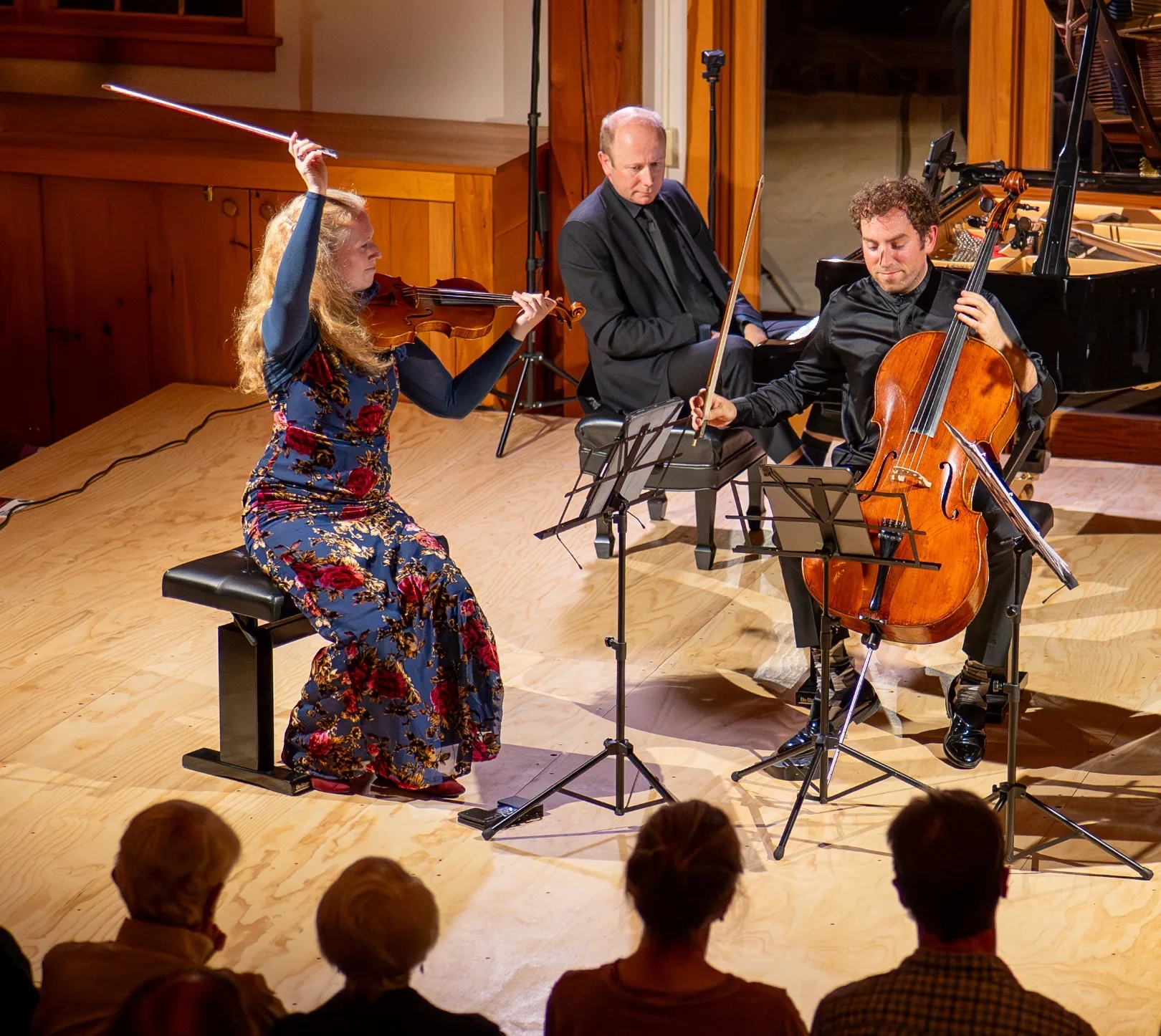 A woman playing violin and a man playing cello perform together on stage, with a pianist in the background. An audience watches them. Music stands and a grand piano are also visible.