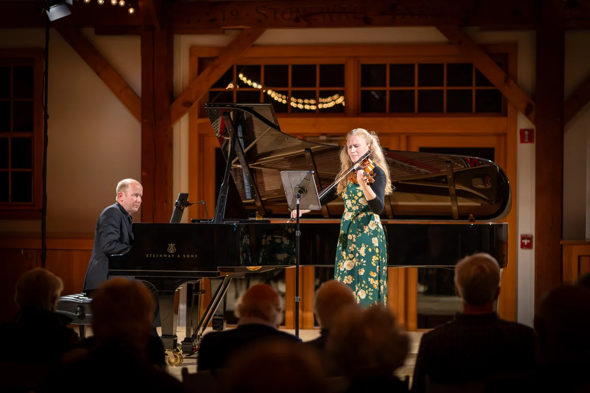 A woman in a green floral dress plays the violin beside a grand piano, accompanied by a pianist, on a warmly lit stage in front of an audience. The setting has wooden beams and large windows.